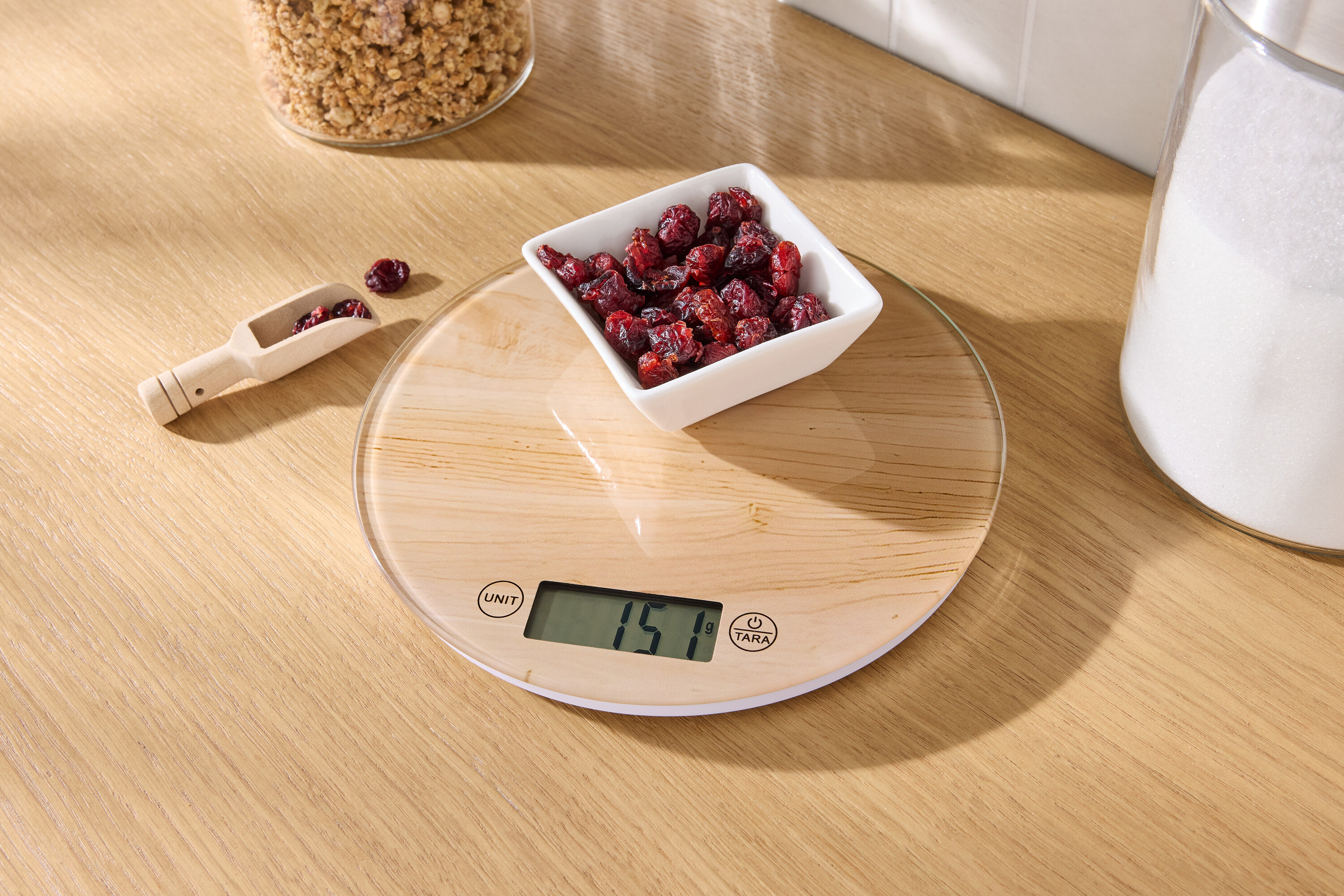 Auf einem Holztisch steht eine Küchenwaage mit einer Schale getrockneter Cranberries und einem hölzernen Messlöffel mit Cranberries.