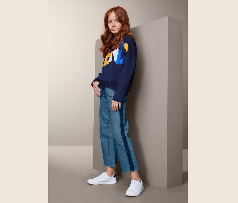 Ein junges Mädchen mit gewellten, roten Haaren posiert in einem dunkelblauen Sweatshirt, einer Jeans-Culotte mit Streifen und weißen Turnschuhen.