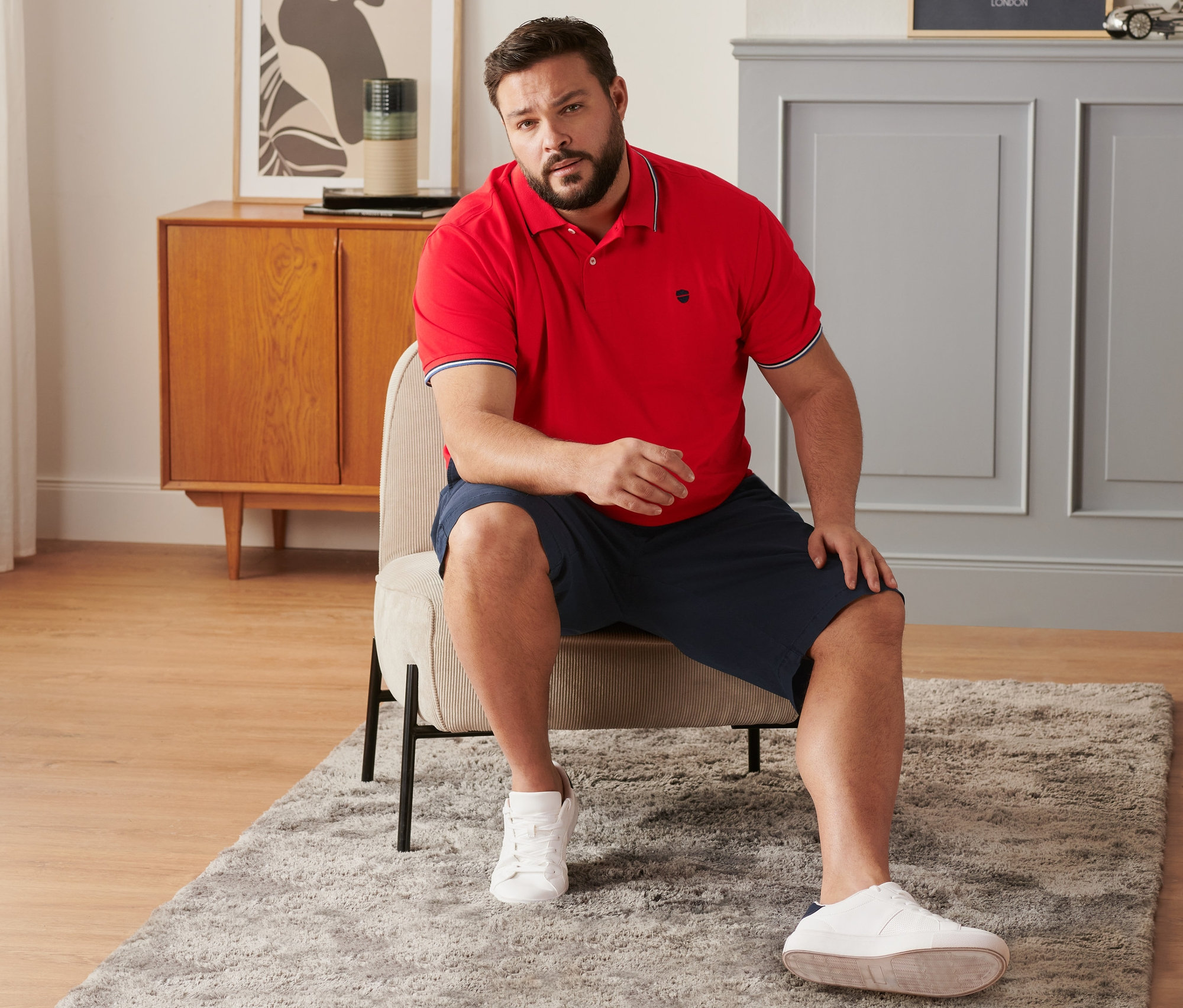 Mann sitzt auf einem Stuhl und trägt marineblaue Chino-Shorts, ein rotes Piqué-Poloshirt und Sneaker.