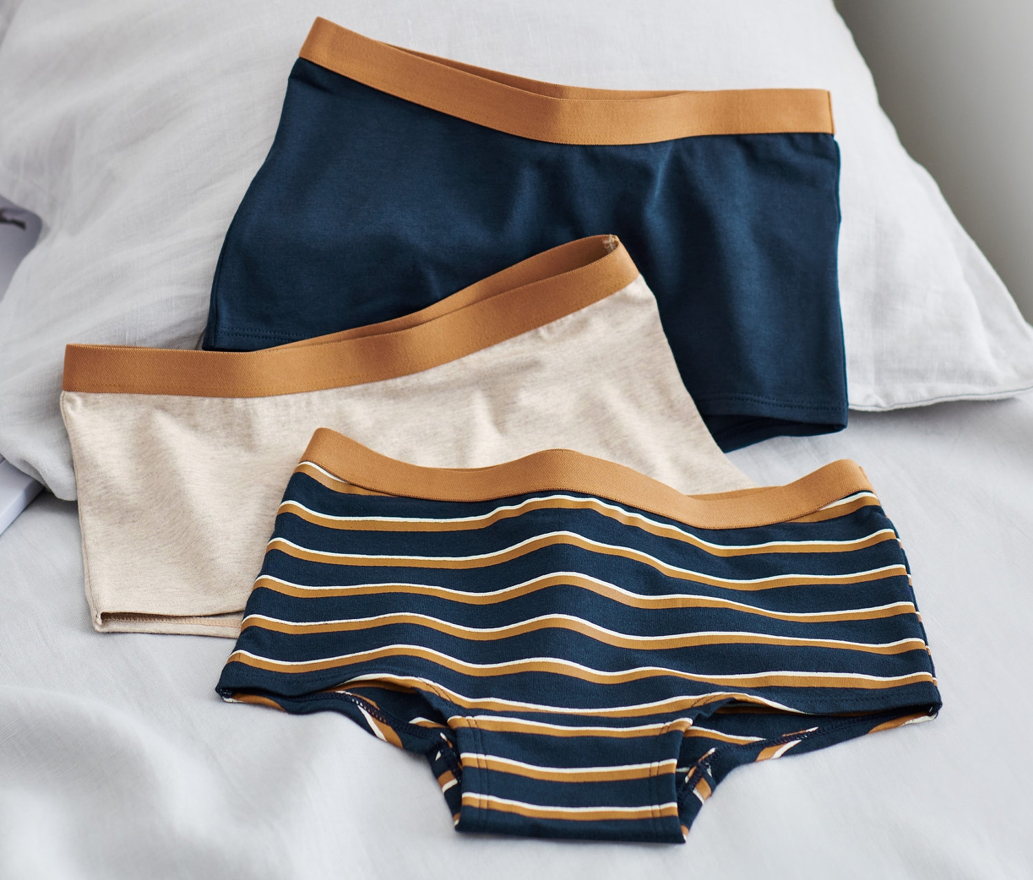 Drei Kinder-Pantys liegen auf einem Bett: eine dunkelblaue, eine beige und eine blau-weiss gestreifte.