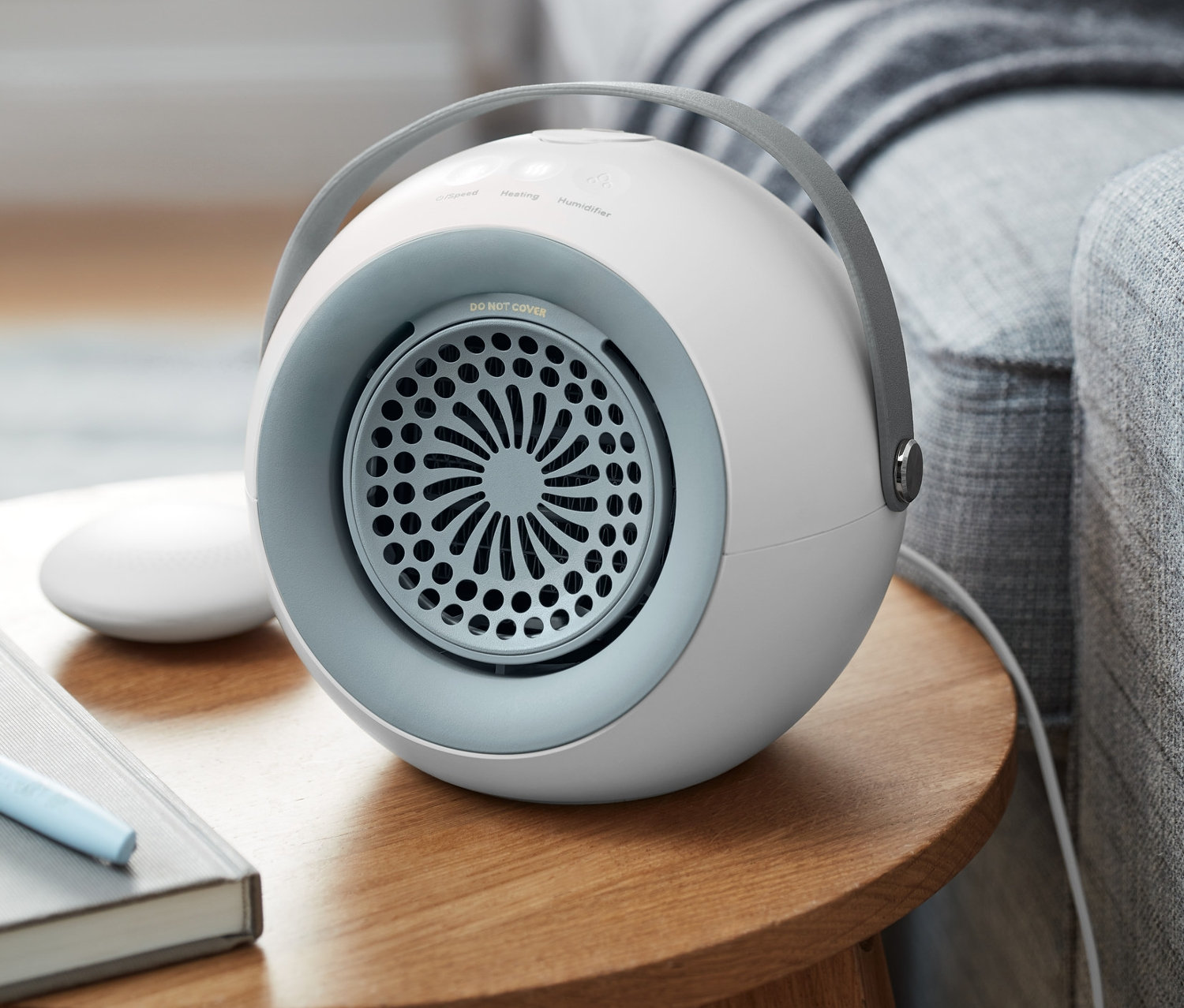 Ein Heizlüfter mit abnehmbarem Handwärmer steht auf einem Holztisch neben einem grauen Sofa.