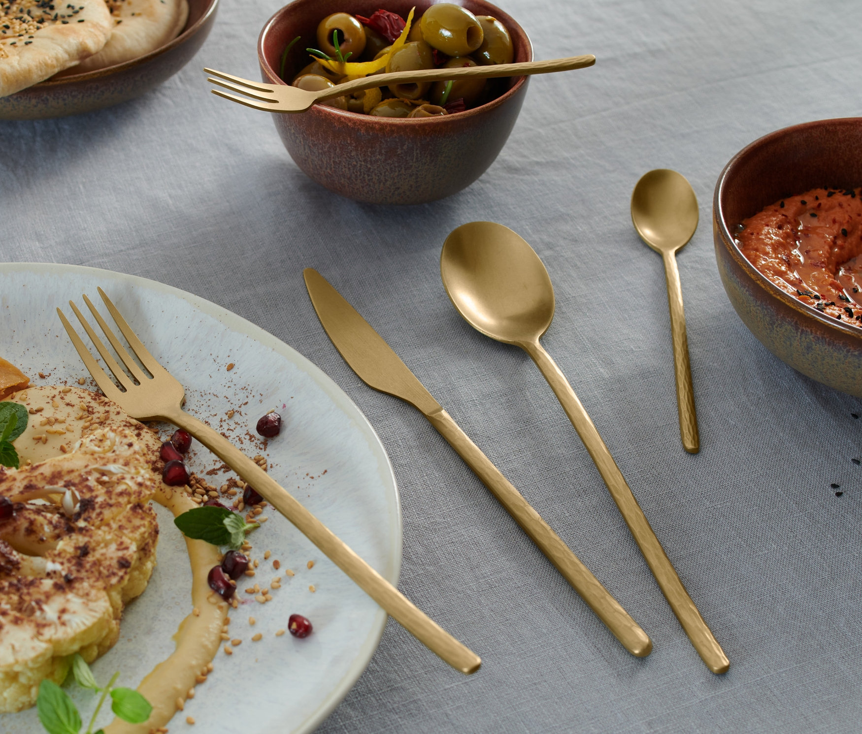 Teller mit Speisen, Schüsseln und ein 20-teiliges Besteck-Set auf einer grauen Tischdecke.