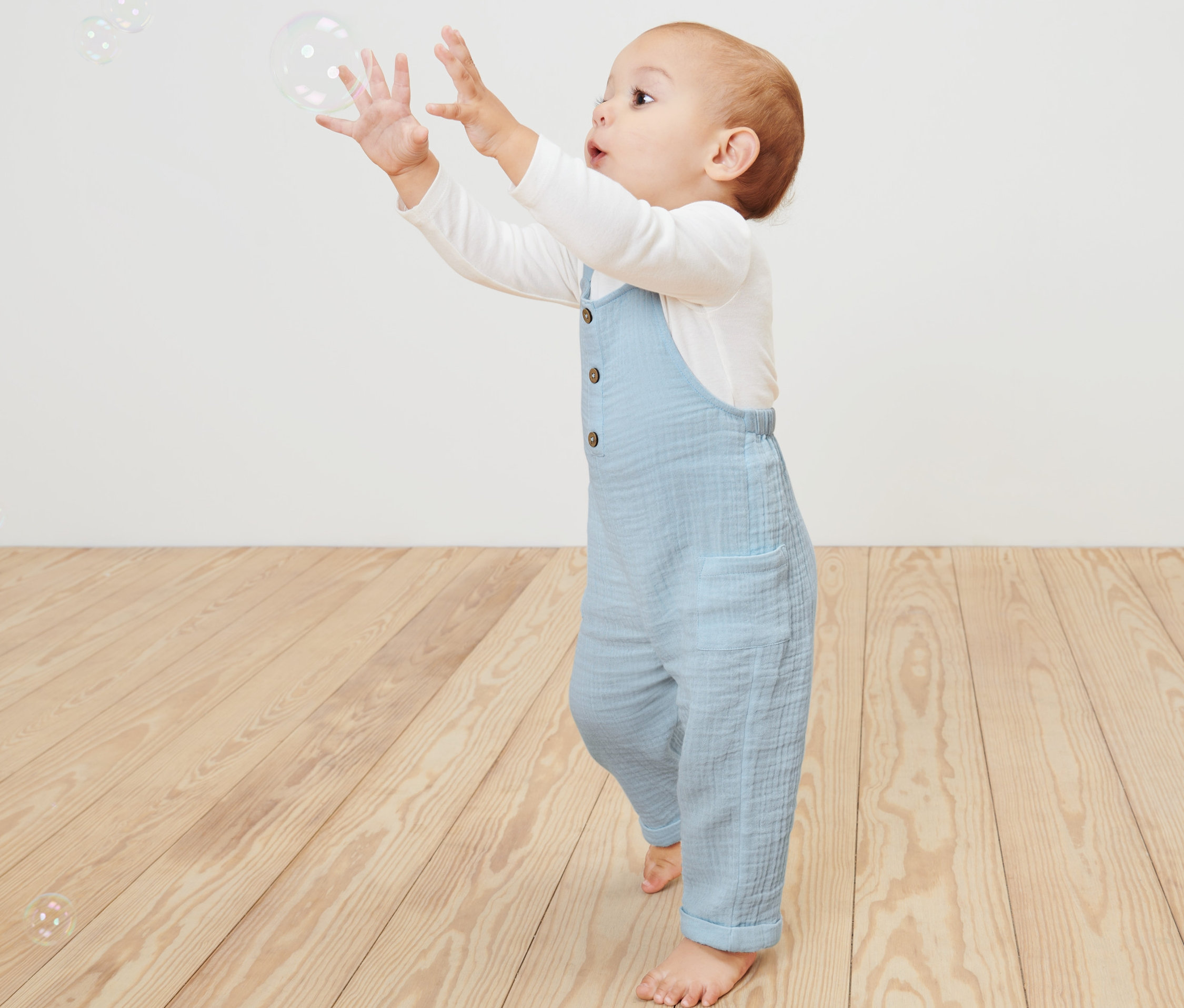 Kleinkind greift nach Seifenblasen in blauer Baby-Latzhose.