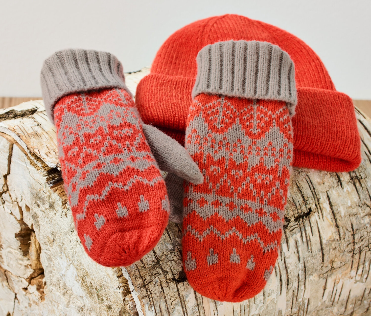 Strick-Fäustlinge und eine orangefarbene Strickmütze liegen auf einem Birkenholzscheit.