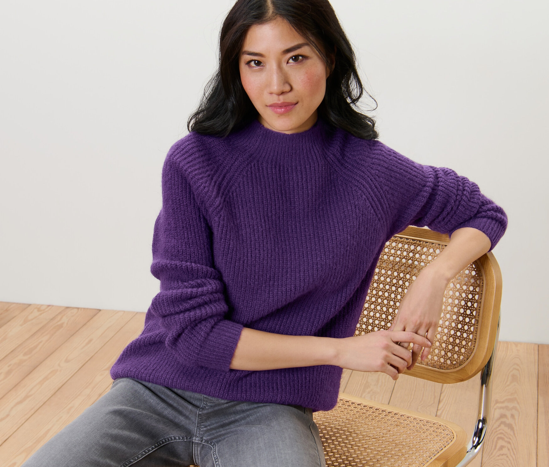 Frau sitzt auf einem Stuhl und trägt einen lila Grobstrickpullover mit Stehkragen.