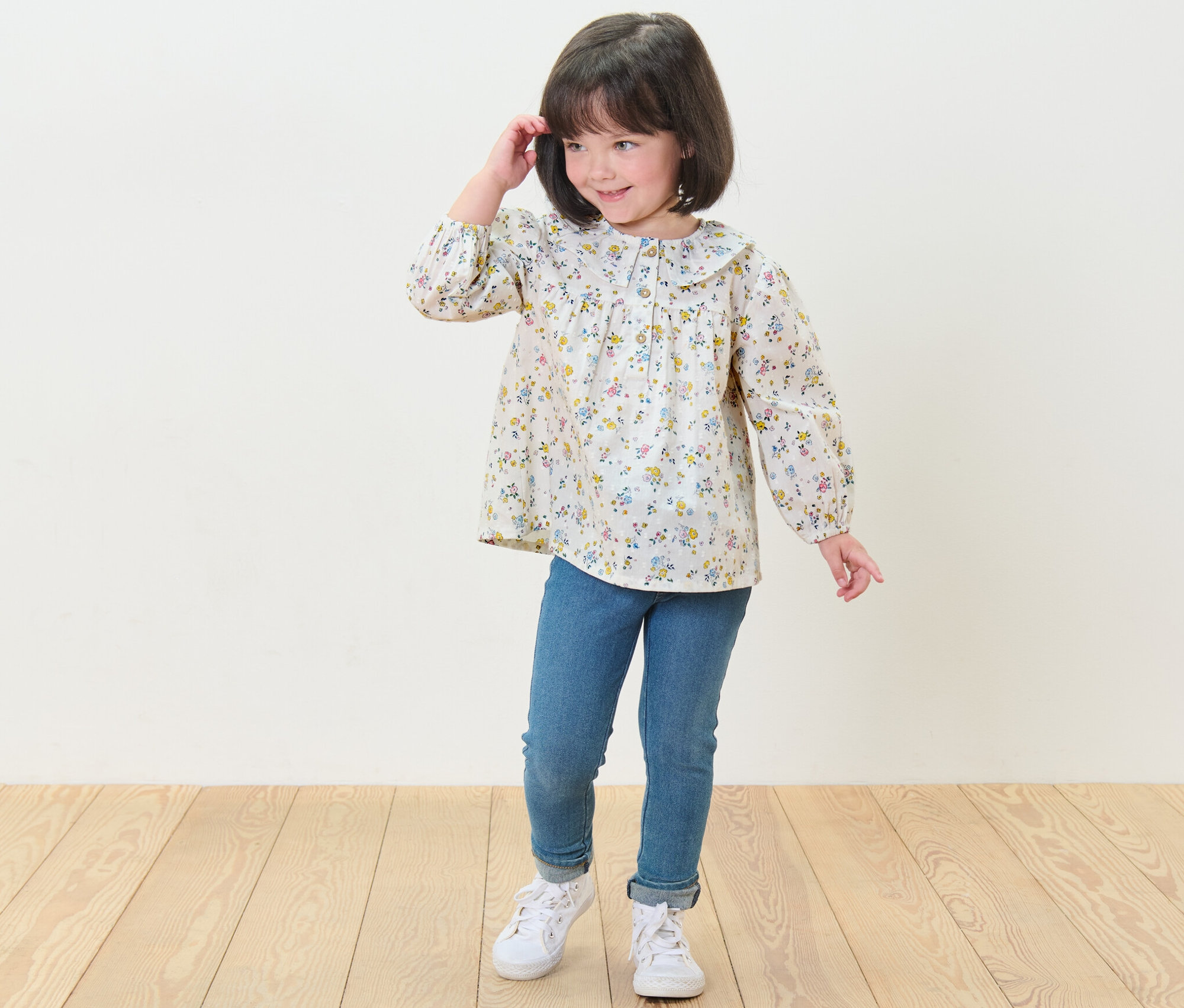 Ein lächelndes Mädchen posiert in einer Kinder-Bluse mit Blumenmuster, blauen Jeans und weißen Turnschuhen.