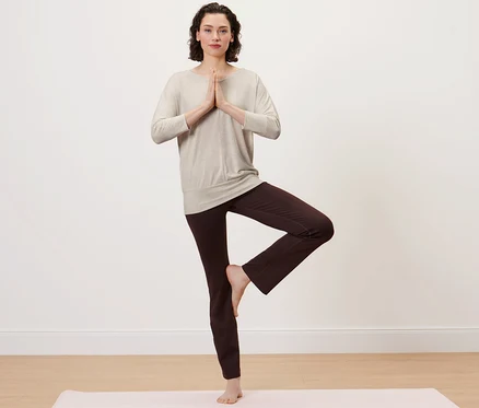 Frau im beigen Yogashirt steht in Baumpose auf einer Yogamatte.