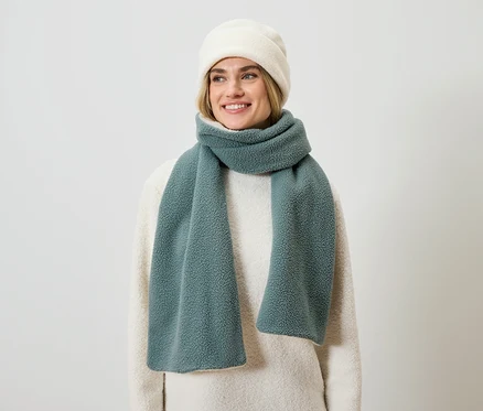 Eine lächelnde blonde Frau trägt einen cremefarbenen Pullover, einen blaugrünen Schal und eine cremefarbene Mütze.