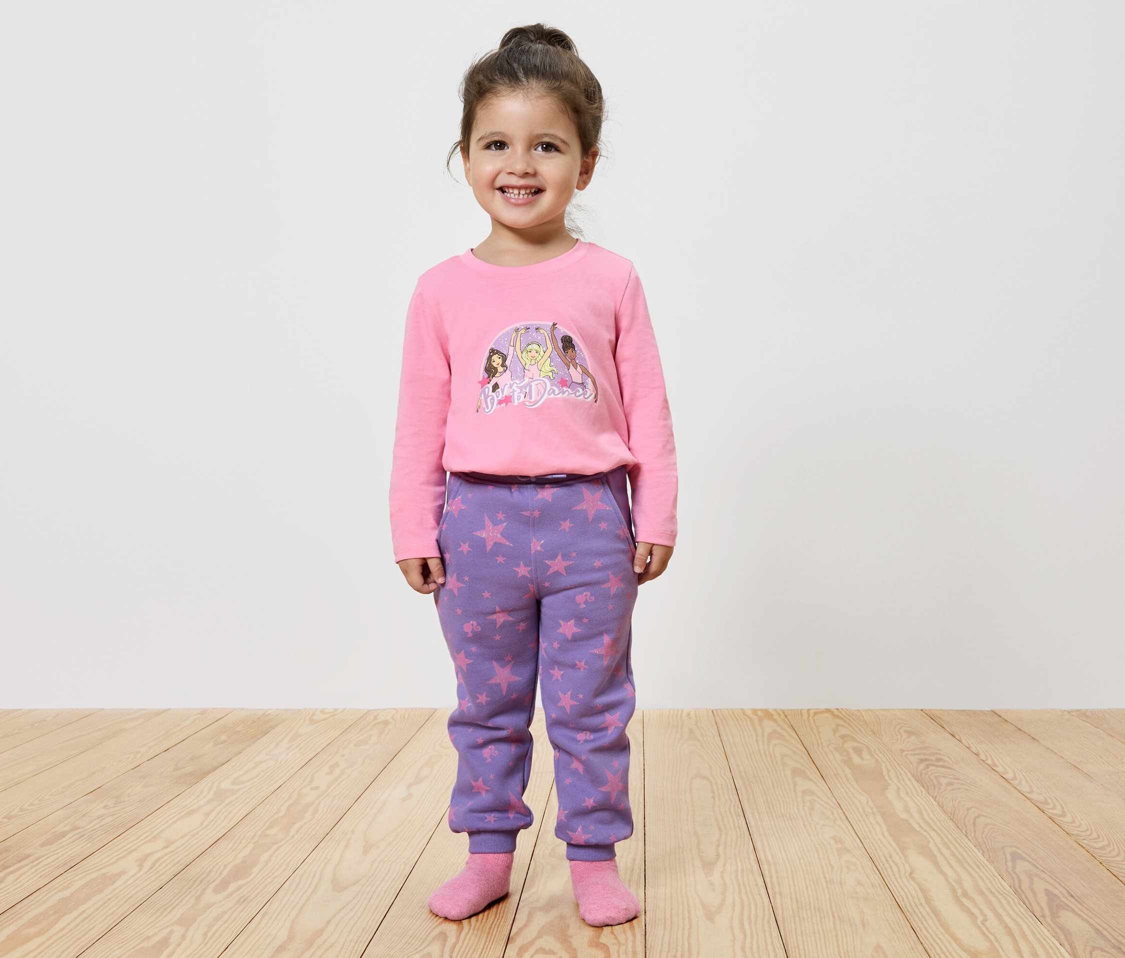 Lächelndes kleines Mädchen posiert in einem rosa bedruckten Langarmshirt und lila Sternen-Hosen.