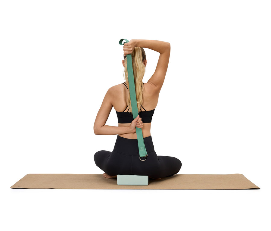 Frau sitzt auf einer Yogamatte mit einem Block darunter und verwendet einen grünen Yogagurt, um ihre Arme hinter dem Rücken zu dehnen.
