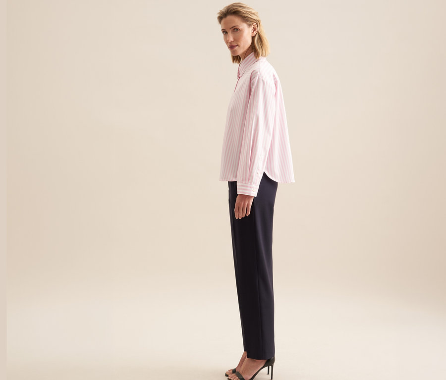 Eine Frau im Profil, den Kopf nach vorne gedreht, trägt eine roséfarbene, gestreifte Seidensticker Oversize Hemdbluse, dunkle Hose und schwarze Sandaletten mit Absatz.