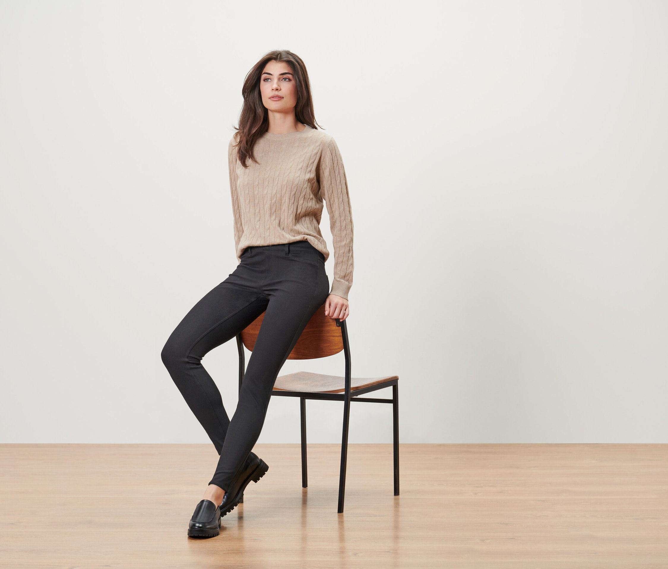 Frau sitzt auf einem Holzstuhl, trägt einen beigen Pullover, schwarze Hosen und schwarze Slipper.