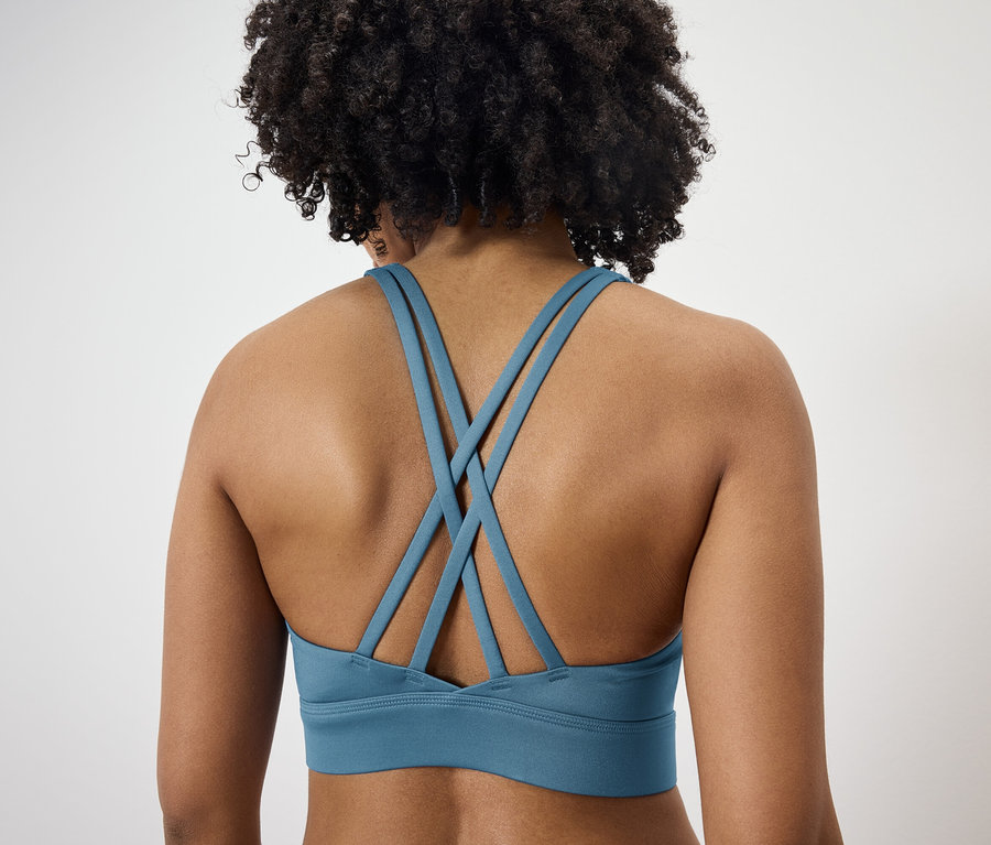 Rückansicht einer Frau mit dunklem lockigem Haar, die einen blauen Sport-BH mit gekreuzten Trägern trägt.