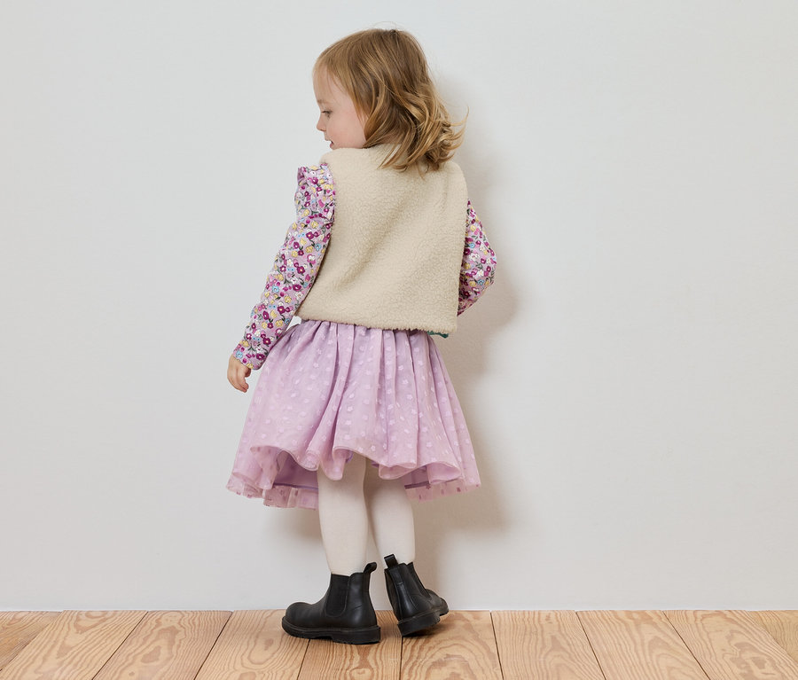 Ein kleines Mädchen steht mit dem Rücken zur Kamera, trägt eine Bluse mit Blumenmuster, eine helle Weste, einen lilafarbenen Rock und schwarze Stiefel.