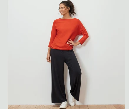 Frau posiert in orangefarbenem Feinstrickpullover, schwarzen Hosen und weißen Turnschuhen.