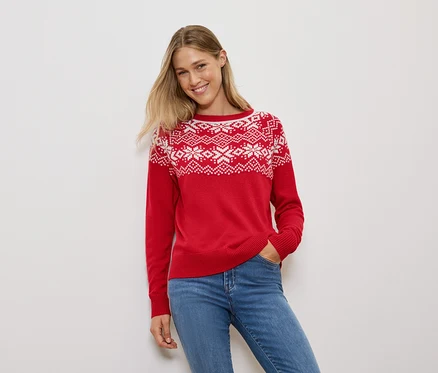Frau mit blonden Haaren posiert in einem roten Pullover mit Schneeflockenmuster und blauen Jeans.