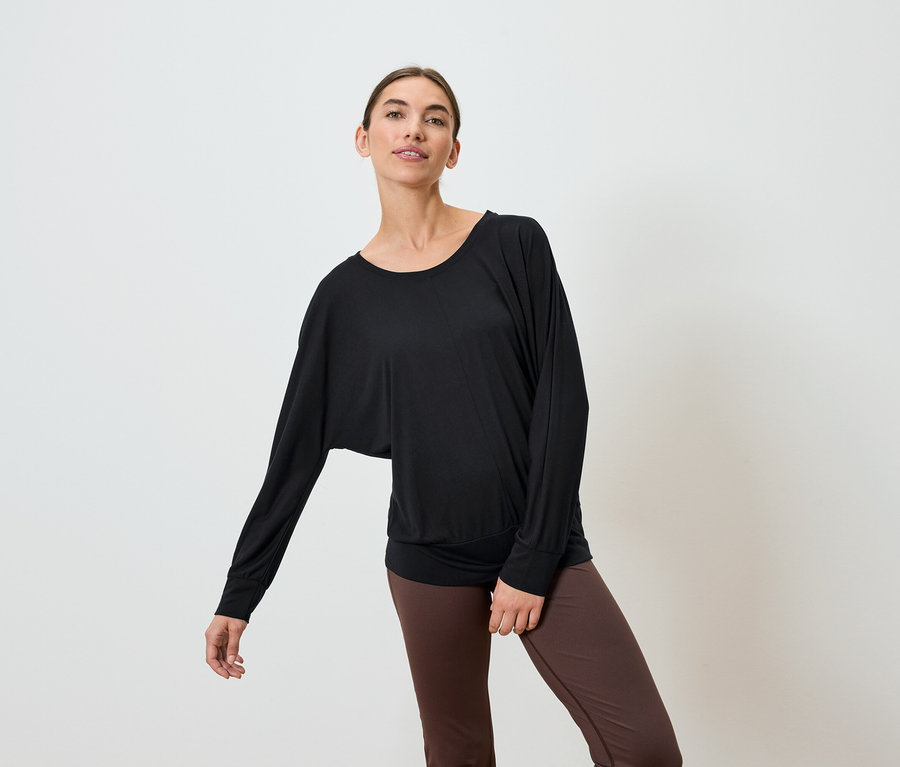 Frau in schwarzem, langärmligem Oberteil und braunen Leggings posiert vor weißem Hintergrund.