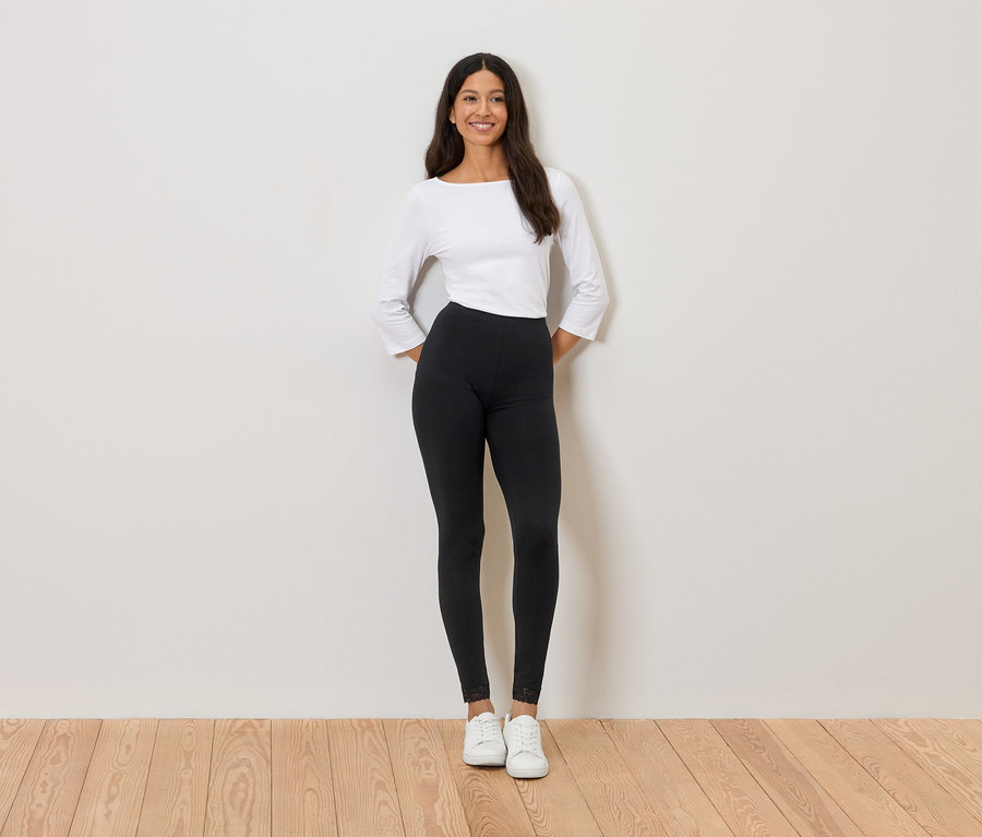 Frau steht vor einer Wand, trägt ein weißes Oberteil, schwarze Leggings und weiße Turnschuhe.