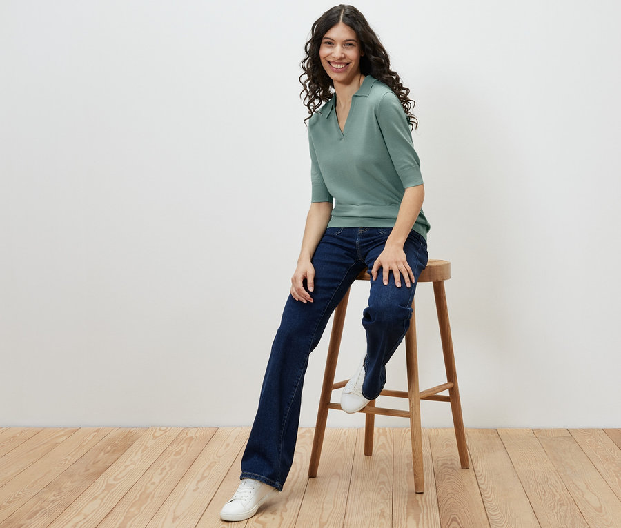 Frau sitzt auf einem Hocker und trägt ein Feinstrick-Polo, Jeans und Turnschuhe.