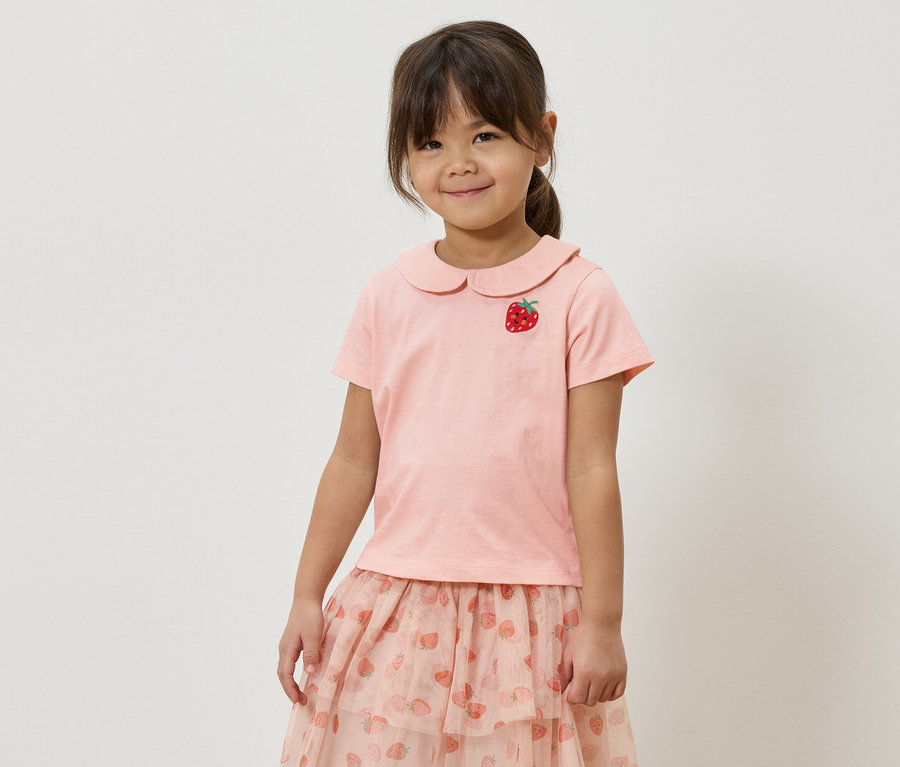 Mädchen mit rosa T-Shirt mit Erdbeer-Applikation und rosa Rock mit Erdbeermuster.