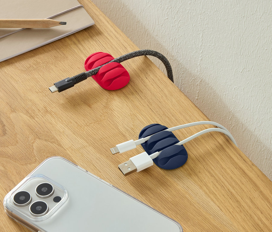 Ein USB-C-Kabel in einer roten Kabelklemme, zwei USB-Kabel in einer blauen Kabelklemme und ein Smartphone auf einem Schreibtisch.