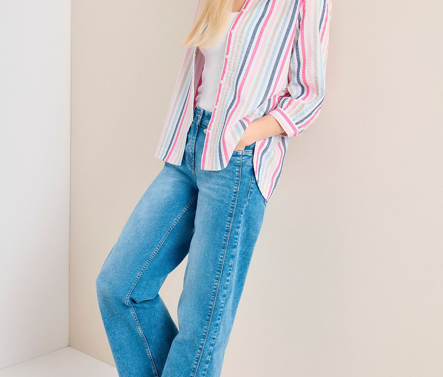 Frau mit hellblauer Kappe, gestreifter CECIL Hemdbluse über weißem Top, blauen Jeans und weißen Sneakern, lehnt an einer Wand.
