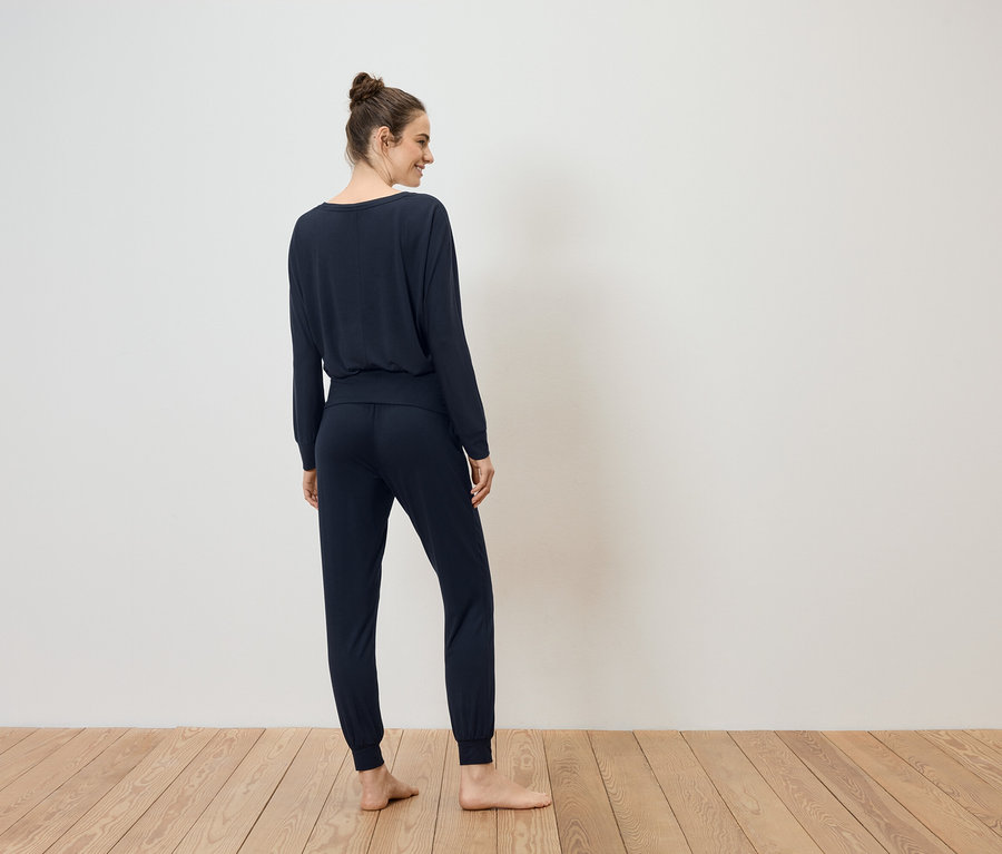 Frau mit Duttfrisur, von hinten in marineblauem, langärmligem Oberteil und Jogginghose auf Holzboden vor weißer Wand.