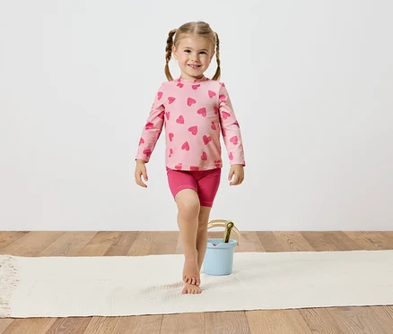 Mädchen mit Zöpfen, trägt ein rosa Herzchenoberteil und Shorts, balanciert auf einem weißen Teppich neben einem hellblauen Eimer.