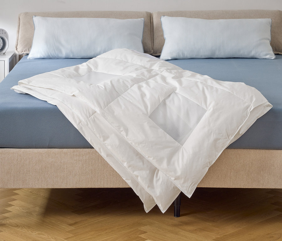 Eine weiße Bettdecke liegt auf einem Bett mit blauem Spannbettlaken und zwei Kissen. Auf dem Nachttisch steht ein Ventilator.