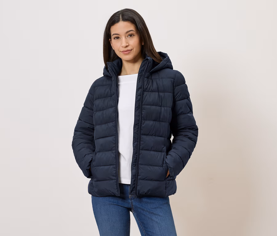 Frau mit marineblauer Steppjacke mit Kapuze, weißem T-Shirt und Jeans, die mit den Händen in den Taschen vor einer weißen Wand steht.