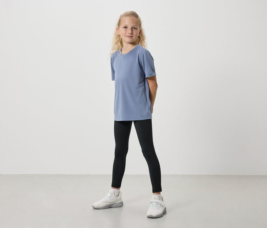 Mädchen steht frontal in blauem T-Shirt, schwarzen Leggings und weißen Turnschuhen vor einem hellen Hintergrund.
