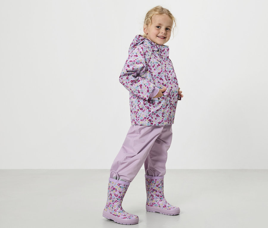 Ein blondes Mädchen trägt einen lila Regenmantel mit Blumenmuster, eine lila Regenhose und passende Gummistiefel.