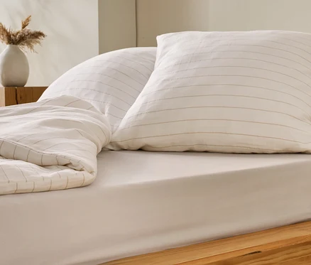 Bett mit beiger Bettwäsche und zwei Kissen mit beigen Streifen. Auf dem Nachttisch steht eine Vase mit Trockenblumen.