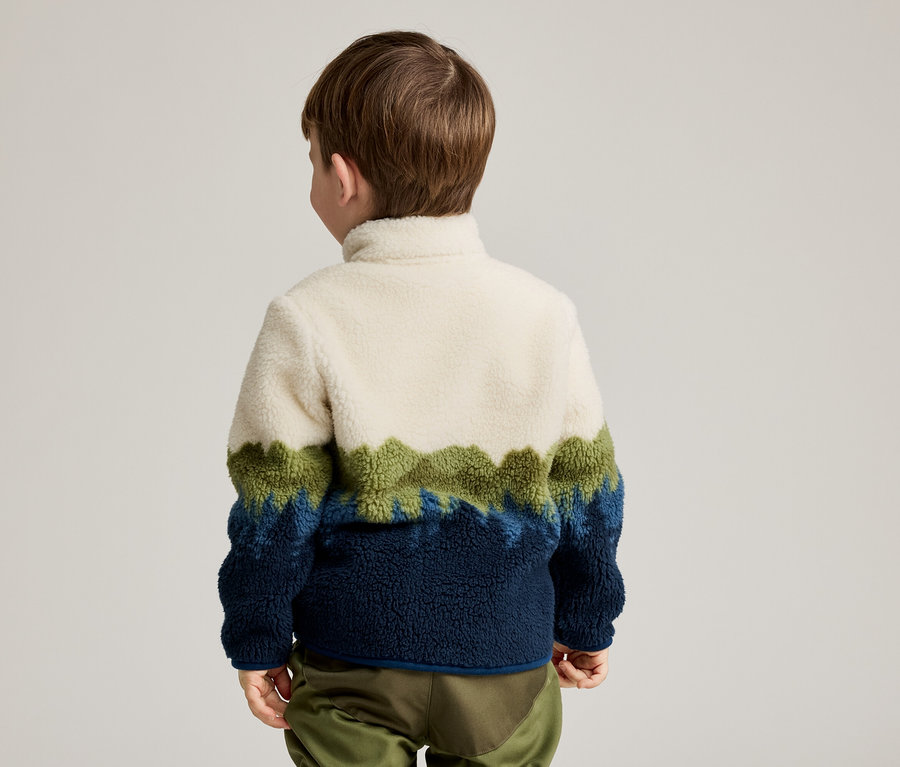 Rückansicht eines Kindes mit braunen Haaren, das einen Fleece-Pullover in Weiß, Grün und Blau sowie eine grüne Hose trägt.