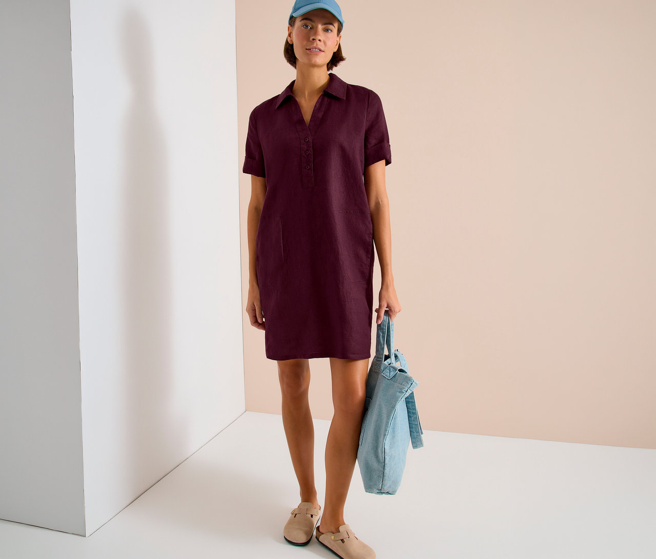 Frau in weinrotem Hemdkleid, blauer Kappe und beigen Clogs. Sie hält eine hellblaue Stofftasche.