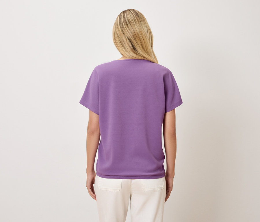 Rückansicht einer Frau mit blonden Haaren, die ein violettes T-Shirt und cremefarbene Hosen trägt.