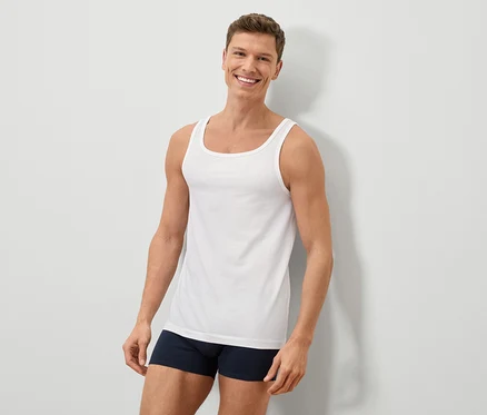 Ein lächelnder Mann mit kurzem braunem Haar trägt ein weißes Tanktop und blaue Boxershorts vor einer weißen Wand.