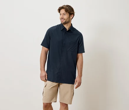 Ein Mann mit Bart trägt ein marineblaues Kurzarmhemd und Khaki-Cargo-Shorts vor einem cremefarbenen Hintergrund.