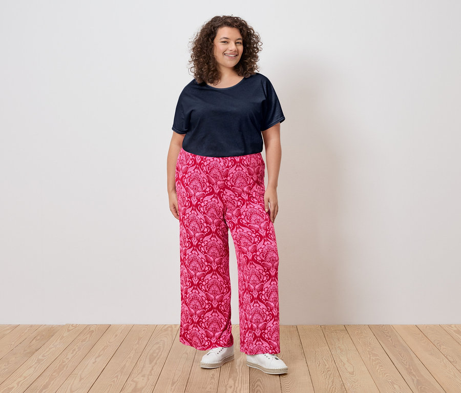 Eine Frau mit lockigem Haar trägt ein marineblaues T-Shirt, eine rosafarbene, gemusterte Hose und weiße Turnschuhe.