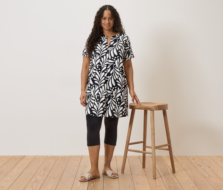 Frau mit lockigem Haar, die ein schwarz-weißes Kleid, Leggings und Sandalen trägt und neben einem Hocker steht.