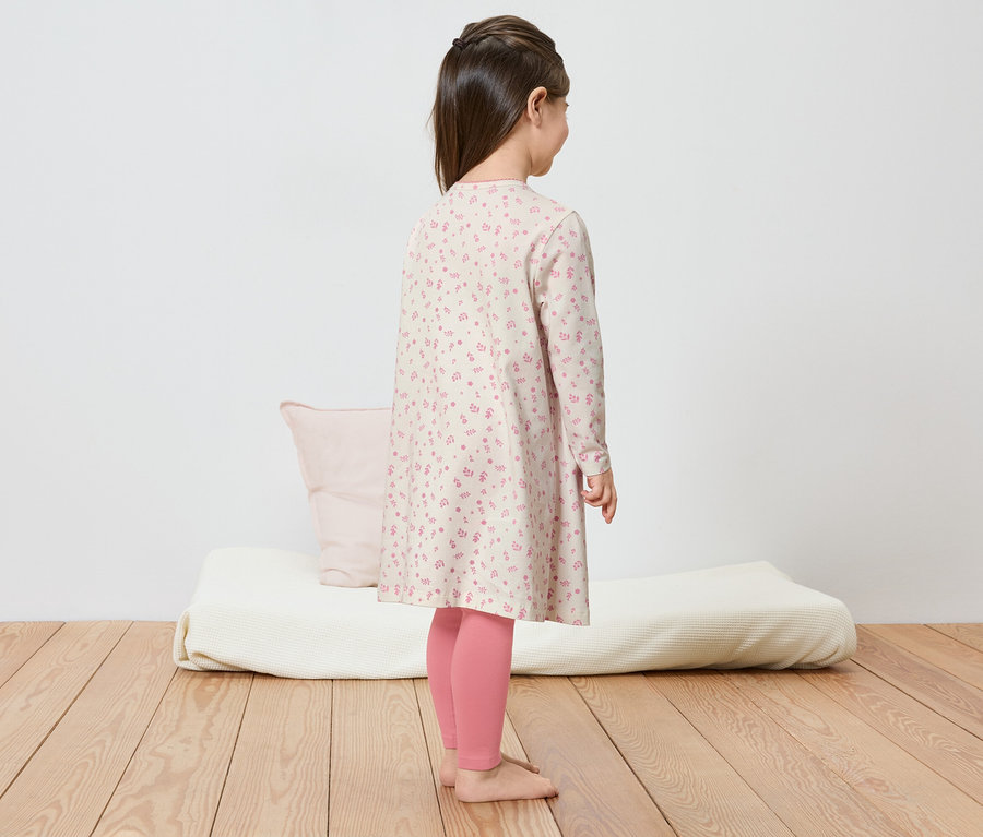 Ein Mädchen mit Zopf steht in einem geblümten Kleid und rosa Leggings auf einem Holzboden.