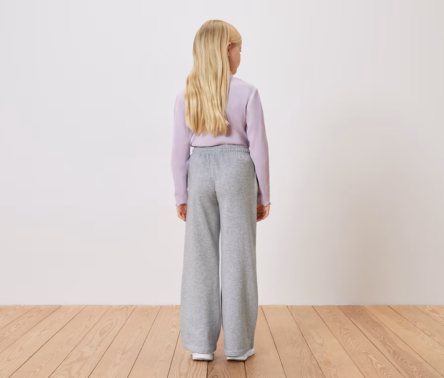 Blick von hinten auf ein blondes Mädchen mit helllila Pullover und grauer, weiter Hose vor einer weißen Wand.