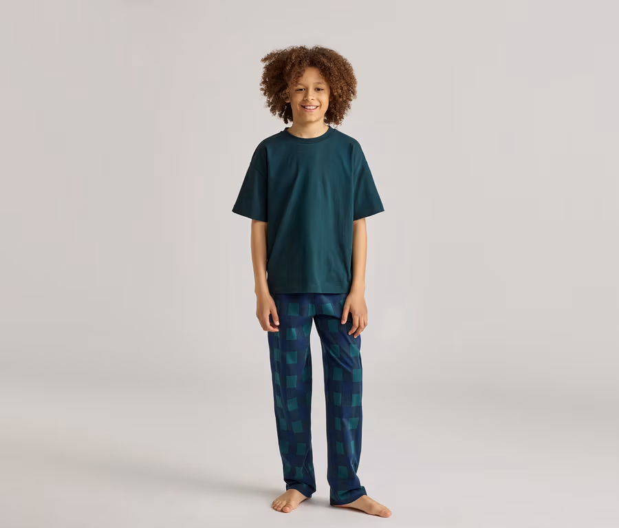 Ein lächelnder Junge mit lockigem Haar trägt einen dunkelgrünen Pyjama mit karierter Hose.