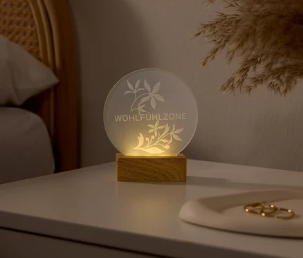 Leuchtende, runde Lampe mit Holzsockel auf einem weißen Nachttisch. Die Lampe zeigt geätzte Blumenmuster und den Schriftzug „WOHLFÜHLZONE“. Im Hintergrund sind ein Rattan-Kopfteil und Pampasgras zu sehen.
