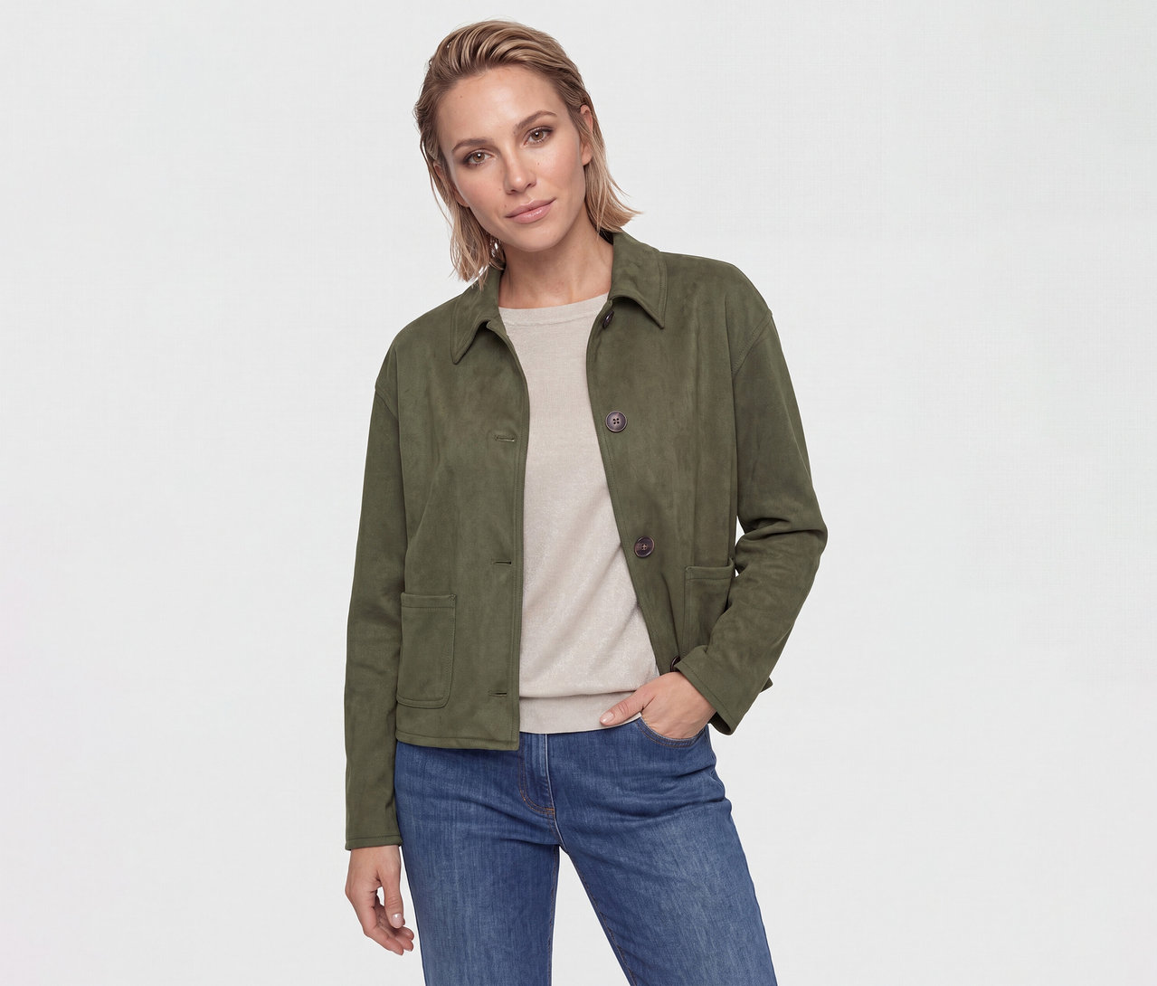Eine Frau mit kurzen, hellbraunen Haaren trägt eine dunkelgrüne Jacke, ein beiges Oberteil und blaue Jeans. Ihre Hand steckt in der Hosentasche.