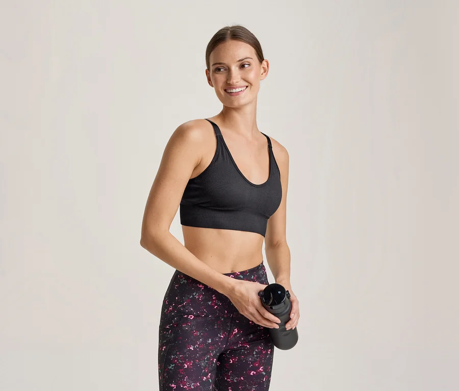 Lächelnde Frau in schwarzem Sport-BH und gemusterten Leggings hält eine schwarze Trinkflasche.