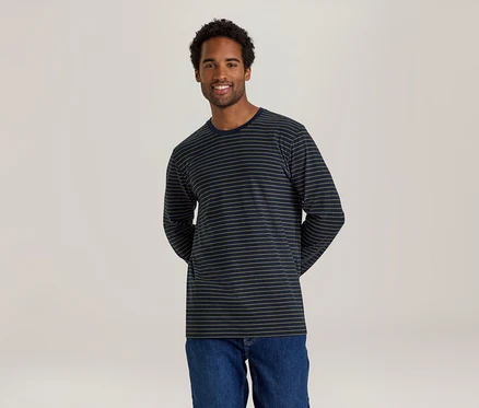Ein lächelnder Mann mit dunklen Locken steht vor einem hellen Hintergrund und trägt ein dunkelblaues Langarmshirt mit dünnen beigen Streifen und Jeans.
