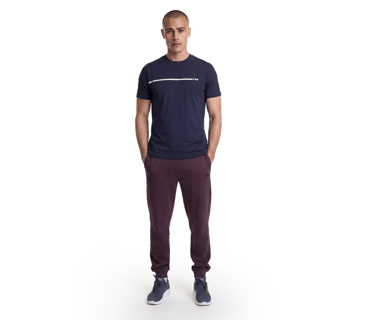Mann steht mit Händen in den Taschen, trägt ein dunkelblaues T-Shirt mit weißem Streifen und Fila-Logo, bordeauxrote Fila-Jogginghose und dunkelblaue Sneaker.