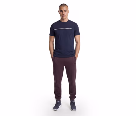 Mann steht mit Händen in den Taschen, trägt ein dunkelblaues T-Shirt mit weißem Streifen und Fila-Logo, bordeauxrote Fila-Jogginghose und dunkelblaue Sneaker.