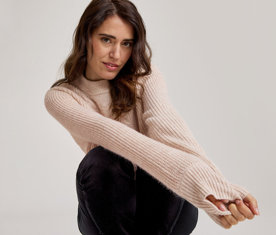 Eine Frau mit langen, dunklen Haaren, die einen hellrosa gerippten Strickpullover mit Daumenlöchern und dunkle Hosen trägt, hockt und blickt in die Kamera.